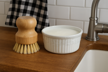 Load image into Gallery viewer, Wooden dish brush and white ceramic bowl on a kitchen counter with a checkered towel and faucet in the background.
